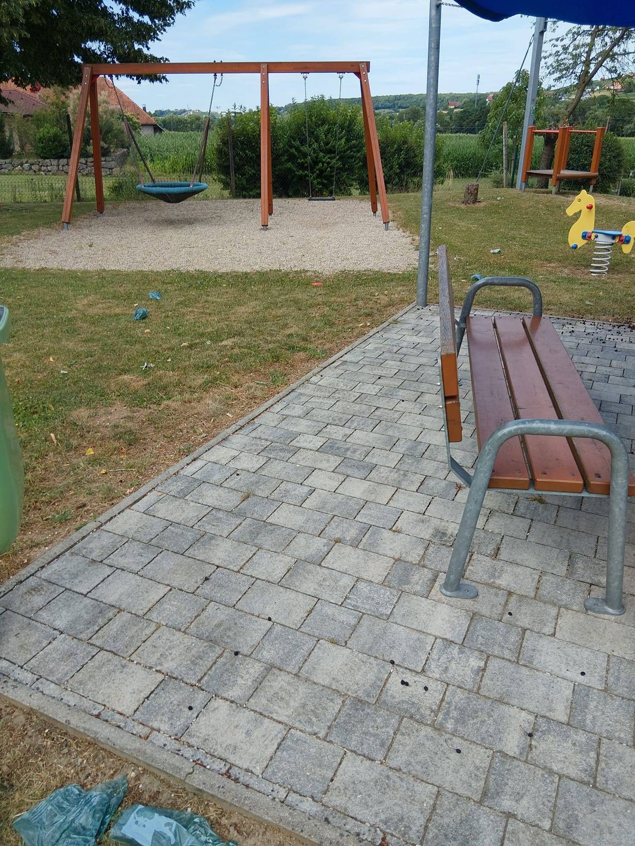 A wooden bench is on a bricked walkway next to a playground with swings and slides.