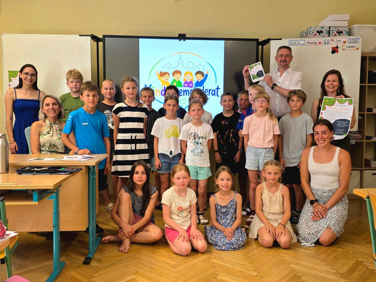 Ein Klassenzimmer mit Schülern und zwei Lehrern, die für ein Gruppenfoto posieren, mit einer Projektionsleinwand im Hintergrund, die ein Bild von Kindern zeigt.