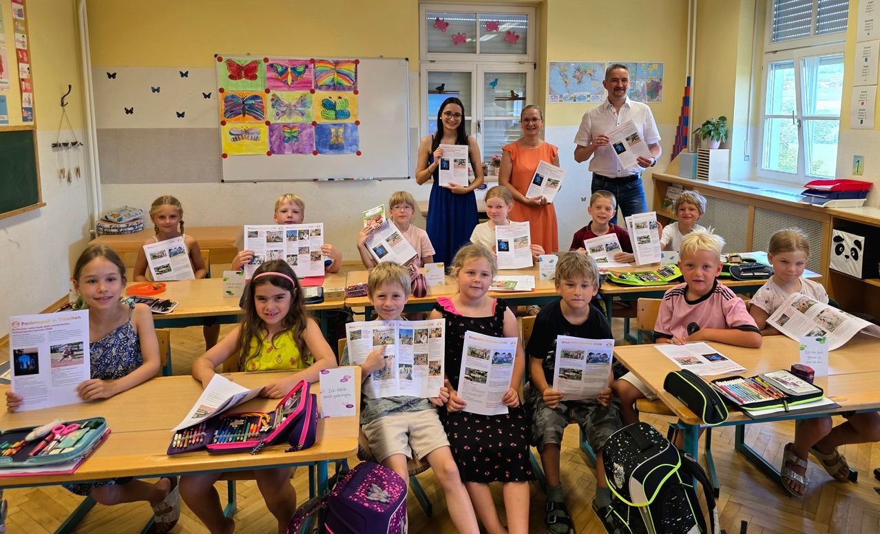A group of children and adults in a classroom hold up colorful magazines and papers. A butterfly collage and world map decorate the walls.