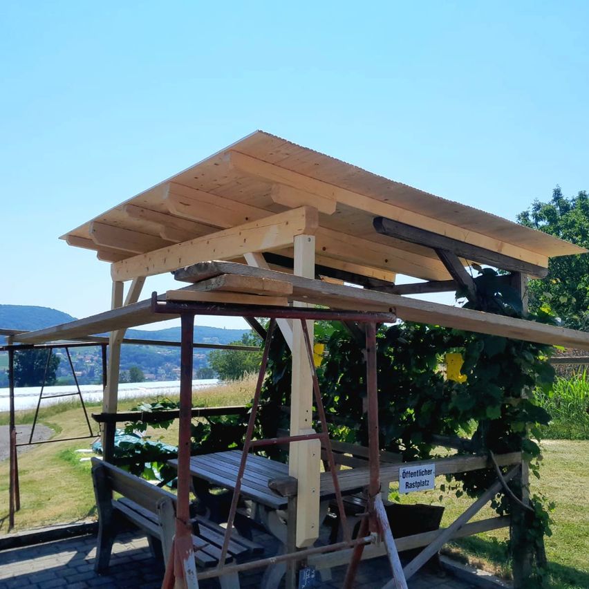 A wooden gazebo with a bench and table under it, with a sign that reads 'Offentlicher Rastplatz'. The gazebo is on a brick floor.