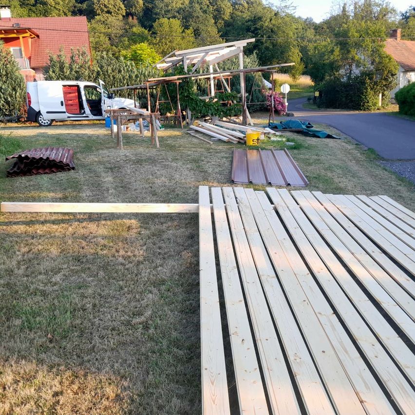 A van is parked in a yard with a wooden structure under construction. Wooden planks and other materials are scattered on the ground. Trees and houses are in the background.
