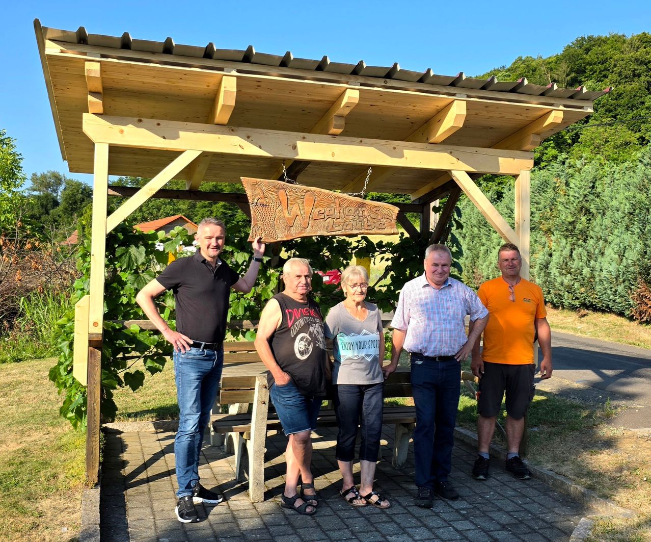 Five people stand under a wooden gazebo with a sign reading 'Weinbergs'. They are smiling for the camera. Behind them, there is a wooden bench and a signboard. Trees and plants are in the background.