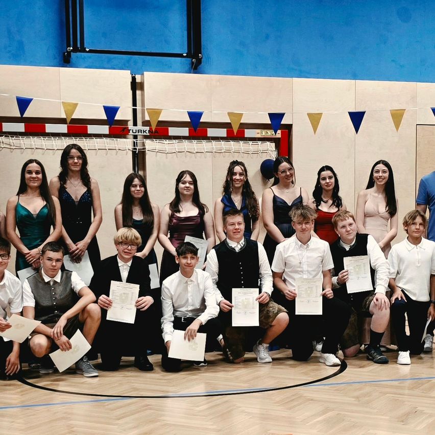 A group of students in formal attire holding certificates pose for a photo in a gymnasium.