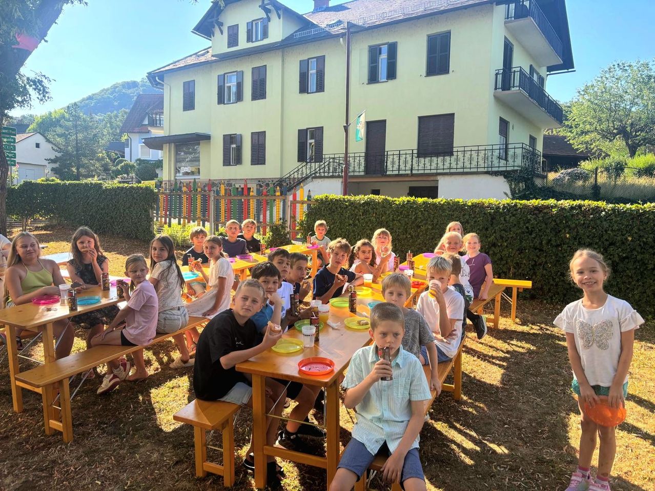 A group of children are sitting at a long table outdoors, eating and drinking. The table is surrounded by a colorful fence. Behind them is a house with many windows and a flag.