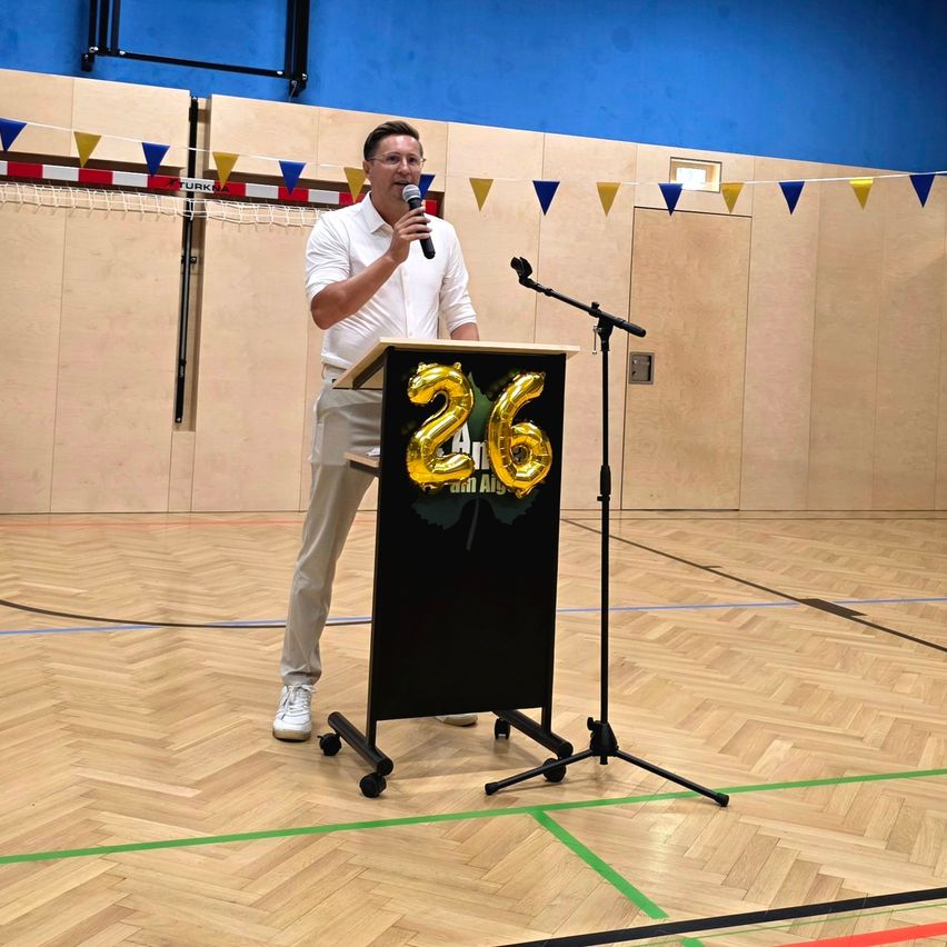 A man stands at a podium with a microphone, speaking into it while a large number 26 is displayed on the podium behind him. The setting is a gymnasium with wooden floors and colorful decorations.