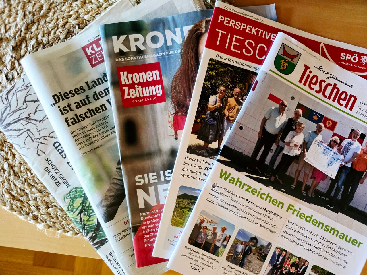 A collection of newspapers on a table, including Kronen Zeitung, Perspektive Tiesc, and Wahrzeichen Frieder. The Kronen Zeitung has a red cover with a blurred person's face.