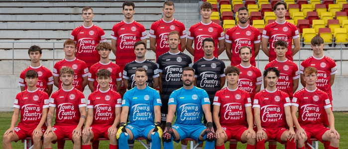 Eine Fußballmannschaft in roten und blauen Trikots posiert für ein Foto auf einem Feld mit mehreren Fußballbällen vor ihnen. Die Spieler sitzen auf Stühlen, und der Hintergrund hat einen verschwommenen Blick auf das Stadion mit leeren Sitzen.