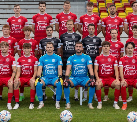 Eine Fußballmannschaft in roten und blauen Uniformen posiert für ein Foto auf dem Feld mit mehreren Fußballbällen. Die Teammitglieder sitzen auf Stühlen.