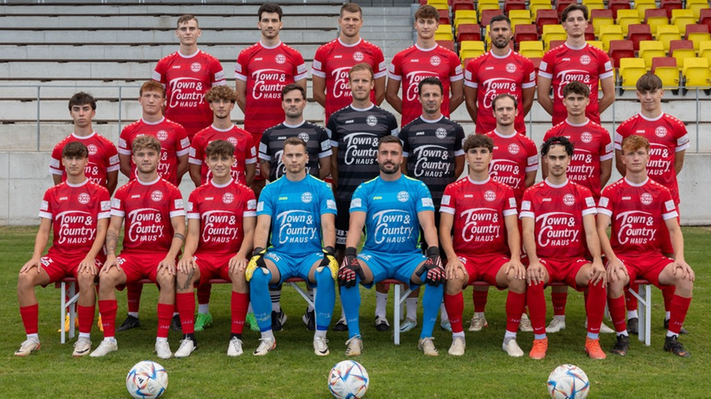Eine Fußballmannschaft in roten und blauen Uniformen posiert für ein Foto auf dem Feld mit mehreren Fußballbällen. Die Teammitglieder sitzen auf Stühlen.