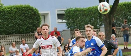 Fußballspieler in roten und blauen Trikots befinden sich auf einem Feld und verfolgen einen Ball in der Luft. Ein Mann ist dabei, den Ball zu treten. Zuschauer schauen von einer Mauer aus zu.