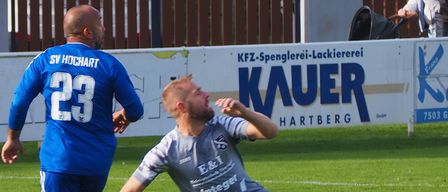 Zwei Fußballspieler auf einem Feld, einer in Blau mit 'SV HOCHART 23' und der andere in Grau, der auf dem Rasen rutscht. Dahinter eine Hecke mit einem Schild für 'KFZ-Spenglerei-Lackiererei' und 'KAUER HARTBARG'.