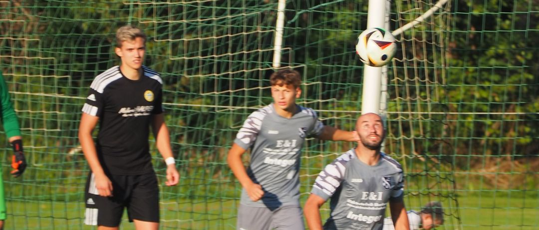 Ein Fußballspiel ist im Gange mit Spielern in schwarzen, grauen und grünen Trikots. Ein Spieler in Schwarz geht, während ein anderer in Grau zum Ball in der Luft läuft.