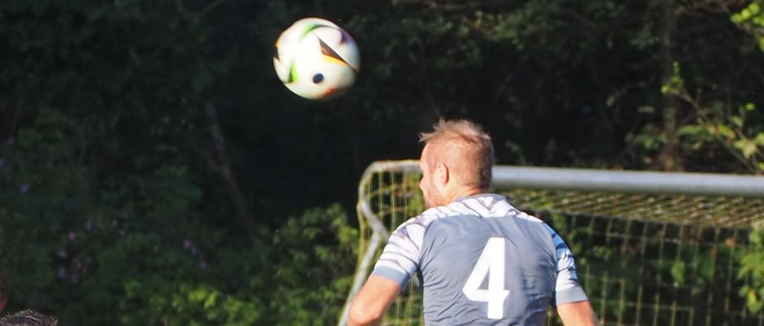 Ein Fußballspieler mit der Trikotnummer 4 springt in die Luft, um den Ball zu fangen. Drei weitere Spieler in schwarzen Trikots stehen hinter ihm.