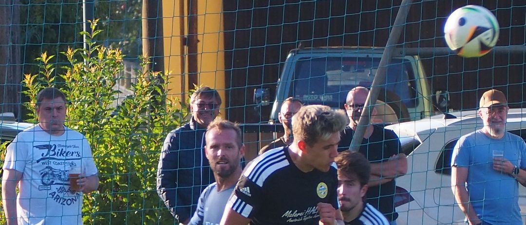 Fußballspieler sind auf einem Feld in Aktion. Ein Spieler ist dabei, den Ball zu schießen. Ein Mann in einem weißen Shirt hält einen Becher. Hinter dem Netz stehen Menschen und schauen zu.