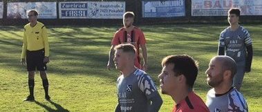 Ein Fußballspiel mit Spielern in roten und grauen Uniformen, einem Schiedsrichter und einem Rasenfeld mit Schatten.