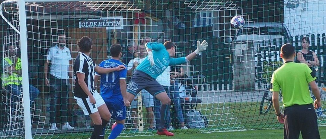 Ein Fußballspieler in einem blauen Trikot versucht, einen Ball, der auf das Tor zufliegt, zu blocken, wobei zwei andere Spieler in der Nähe sind.