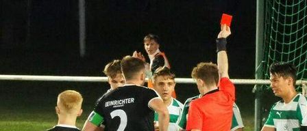 Ein Nachtfußballspiel mit Spielern in grünen und weißen Trikots, ein Spieler liegt auf dem Boden und der Schiedsrichter zeigt einem anderen Spieler eine rote Karte.