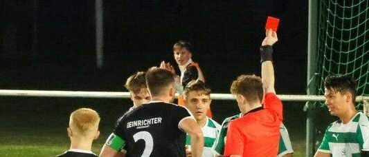 Ein Nachtfußballspiel mit Spielern in grünen und weißen Trikots, ein Spieler liegt auf dem Boden und der Schiedsrichter zeigt einem anderen Spieler eine rote Karte.