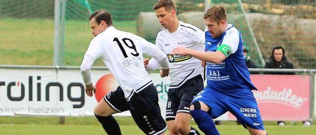 Drei Fußballspieler sind auf einem Feld. Spieler 19 versucht, den Ball zu bekommen, während zwei andere verteidigen. Im Hintergrund befinden sich Werbung auf Bannern.