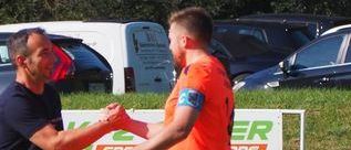 Zwei Männer schütteln sich auf einem Fußballfeld die Hände, mit geparkten Autos und einem Banner im Hintergrund. Ein Mann trägt ein orangefarbenes Trikot, der andere ein schwarzes Hemd.