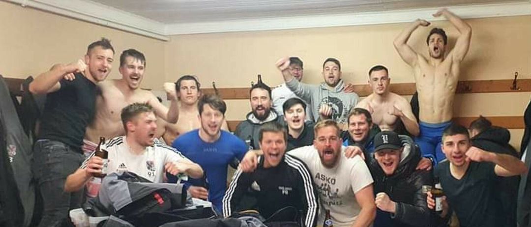 Eine Gruppe junger Männer in einem Raum, einige ohne Hemd, posiert für ein Foto. Zwei halten Bier in der Hand, einer trägt einen Hut. Ein Tisch mit Bierkisten steht hinter ihnen.