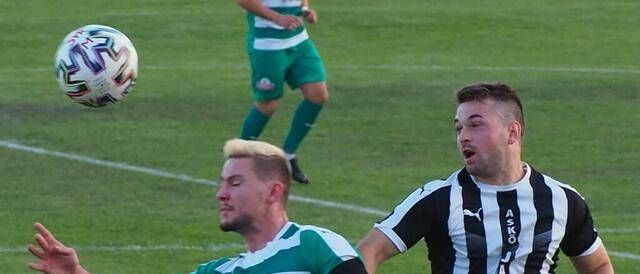 Zwei Fußballspieler konkurrieren auf einem Feld. Ein Spieler in einem grün-weiß gestreiften Trikot greift nach dem Ball, während ein anderer in einem schwarz-weißen Trikot aufmerksam zusieht.