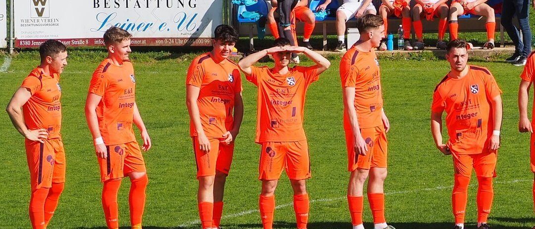 Fußballspieler in orangefarbenen Trikots stehen auf dem Spielfeld. Dahinter sitzen mehrere Spieler in weißen Trikots auf einer Bank. Ein Mann in einem schwarzen T-Shirt geht an der Bank vorbei. Ein Werbeschild ist im Hintergrund zu sehen.