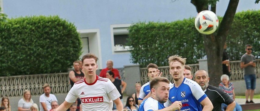 Fußballspieler konkurrieren auf einem Feld, ein Spieler in Rot versucht den Ball zu treten, während ein anderer in Blau verteidigt. Zuschauer beobachten von den Seitenlinien.