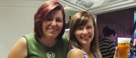 Zwei Frauen lächeln und posieren für ein Foto. Eine trägt ein grünes Shirt mit 'Don't Be The Same' und eine Halskette. Die andere trägt eine Overall-Jeans. Ein Mann in einem gestreiften Hemd ist im Hintergrund verschwommen.