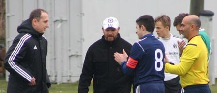 Zwei Männer in Jacken unterhalten sich mit einem Spieler in einer blauen Uniform mit der Nummer 8. Ein Fußball liegt auf dem Rasen.