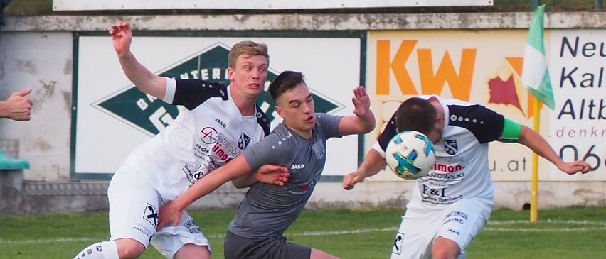 Fußballspieler sind auf einem Feld in Aktion, einer in Weiß und einer in Grau, beide kämpfen um den Ball. Im Hintergrund ein Schild mit dem Wort Car Studio.