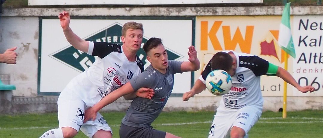 Fußballspieler sind auf einem Feld in Aktion, einer in Weiß und einer in Grau, beide kämpfen um den Ball. Im Hintergrund ein Schild mit dem Wort Car Studio.