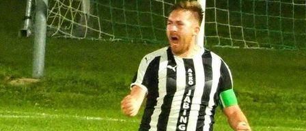 A soccer player in a black and white striped jersey is celebrating on the field with a goal in the background.