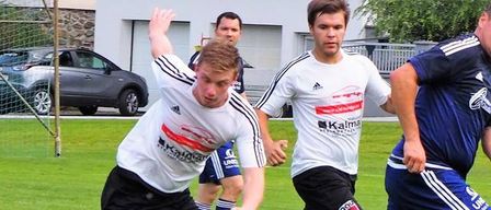 Vier Fußballspieler spielen auf einem Feld. Ein Spieler ist dabei, den Ball zu schießen, während ein anderer Spieler versucht, ihn zu blockieren. Zwei weitere Spieler sind hinter ihnen. Im Hintergrund ist ein Auto vor einem Gebäude geparkt.