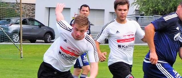 Vier Fußballspieler spielen auf einem Feld. Ein Spieler ist dabei, den Ball zu schießen, während ein anderer Spieler versucht, ihn zu blockieren. Zwei weitere Spieler sind hinter ihnen. Im Hintergrund ist ein Auto vor einem Gebäude geparkt.