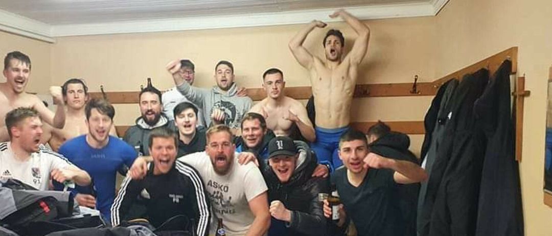 Eine Gruppe junger Männer, einige oberkörperfrei, stehen und sitzen in einem Raum. Ein Mann steht mit erhobenen Armen auf einem Tisch. Sie posieren für ein Foto mit Getränken in der Hand.