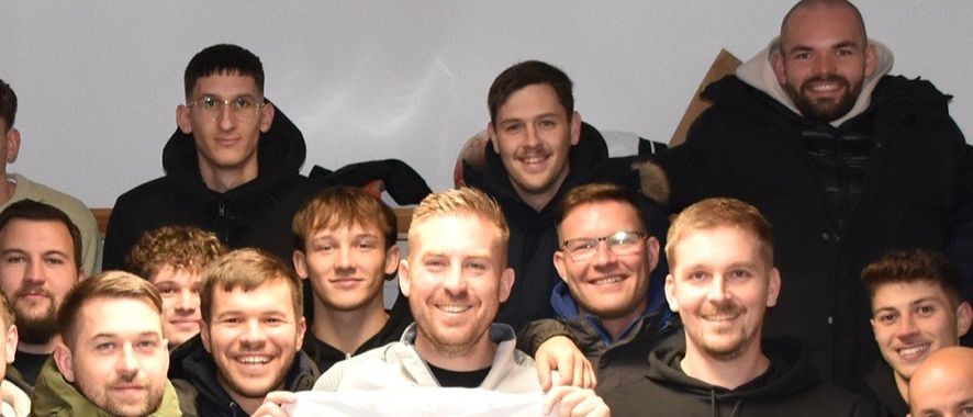 Eine Gruppe von Menschen, darunter Männer in Jacken und Mützen, posiert für ein Foto. Sie halten weiße T-Shirts, auf denen 'Herbstmeister Asko Jabing' steht. Die Shirts haben ein Logo und schwarzen Text. Das Foto hat einen roten Hintergrund mit weißem Text.