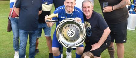 Eine Gruppe von Männern und einem Mädchen posiert mit einem Fußballpokal auf einer Rasenfläche. Ein Mann hält einen goldenen Schuh und den Pokal und lächelt.