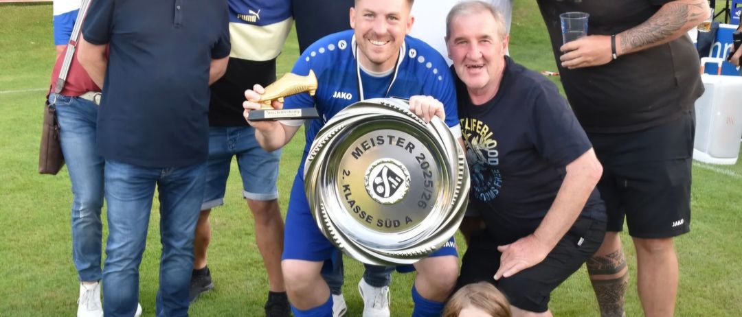 Eine Gruppe von Männern und einem Mädchen posiert mit einem Fußballpokal auf einer Rasenfläche. Ein Mann hält einen goldenen Schuh und den Pokal und lächelt.