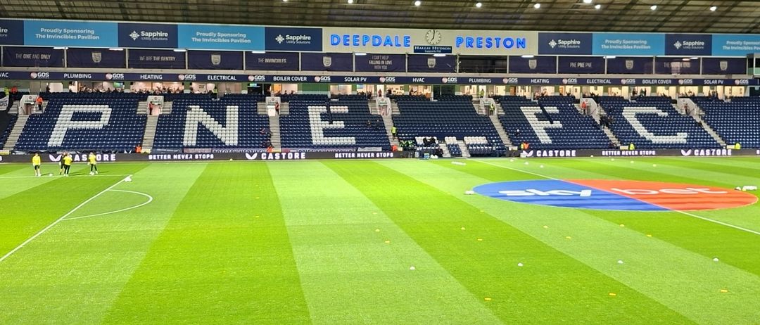 Ein leeres Fußballstadion mit blauen Sitzen, grünem Feld und weißen Linien. Sponsoren wie Castole und Sapphire sind auf Bannern sichtbar. Die Ränge buchstabieren 'INE FC'.
