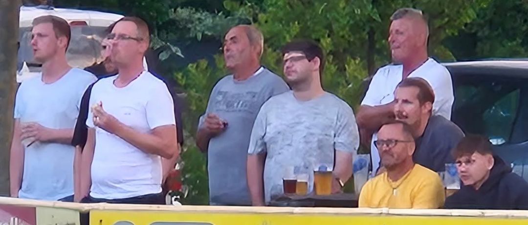 Eine Gruppe von Männern steht hinter einem gelben Banner. Zwei Männer halten Getränke in den Händen. Ein anderer Mann trägt einen Verband auf der Stirn. Das Banner trägt die Aufschrift 'TROC RUPO B202 GRAFE'.