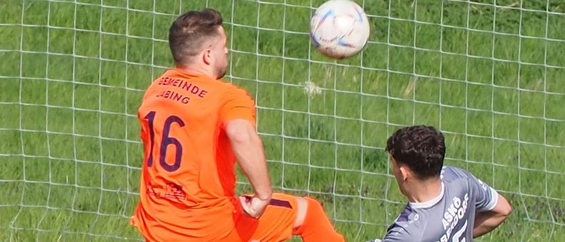 Zwei Fußballspieler, einer in Orange und einer in Grau, sind in ein Spiel verwickelt. Der Spieler in Orange springt, um den Ball zu köpfen, während der Spieler in Grau versucht, ihn zu blockieren.