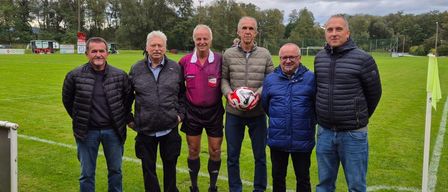 Fünf Männer stehen lächelnd auf einem Fußballfeld. Einer hält einen Fußball. Bäume und ein bewölkter Himmel sind im Hintergrund.