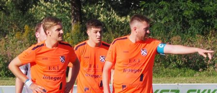 Four soccer players in orange uniforms stand on a field. One player points to the ball while the others watch. A grassy field with a white banner in the background.