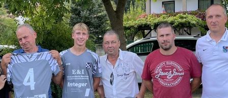 Eine Gruppe von Männern und einer Frau steht in einem Grasbereich mit einem Hund. Ein Mann hält ein graues Trikot mit der Nummer 4.