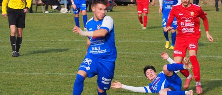 Fußballspieler in blauen und roten Trikots spielen auf einem Feld. Ein Spieler in Blau steht kurz davor, den Ball zu schießen. Dahinter sitzen Leute auf einer Bank und beobachten das Spiel. Ein weißer Wagen ist im Hintergrund geparkt.