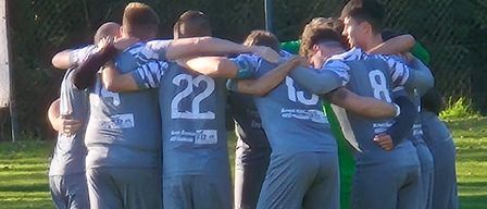 A soccer team huddles together on a grassy field, wearing matching gray uniforms with numbers 22, 10, and 8. Trees and a fence in the background.