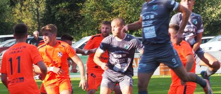 A soccer match is in action on a field, with players in orange and gray uniforms. A player in gray kicks the ball, while another in orange defends. The background shows a hedge and parked cars.