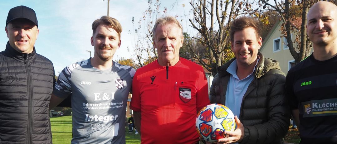 Five men stand on a soccer field. One man holds a soccer ball. Another man wears a referee's red shirt.