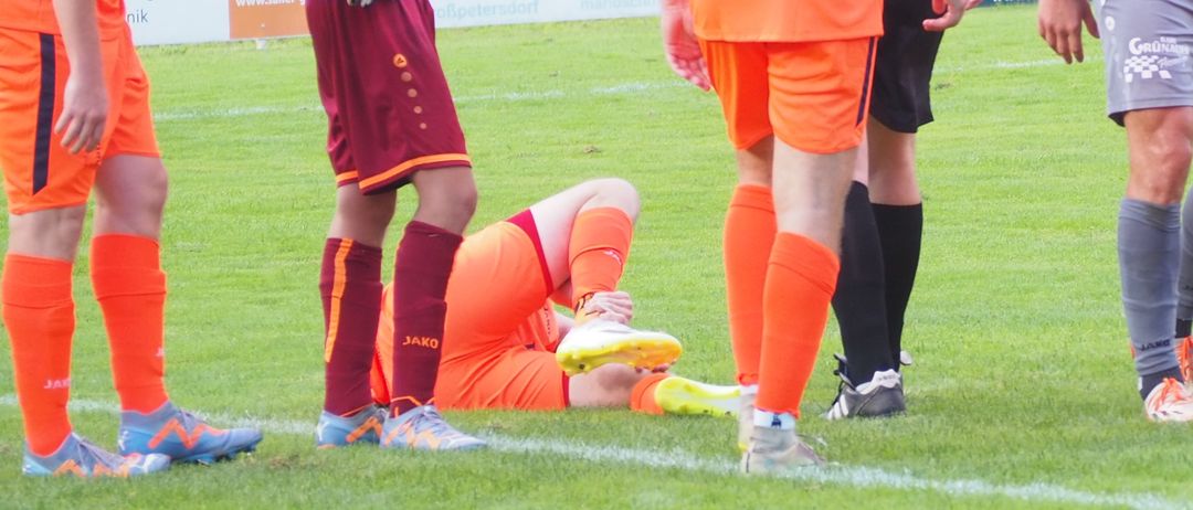 Ein Fußballspiel auf einem Feld mit Spielern in orangefarbenen und grauen Trikots. Ein Spieler liegt auf dem Boden, während andere um ihn herumstehen. Ein Schiedsrichter ist anwesend.